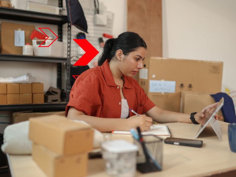 Profissional registra informações manualmente enquanto consulta dados em dispositivo eletrônico, representando processos manuais que aumentam o risco de erros, retrabalho e perda de produtividade na gestão empresarial.